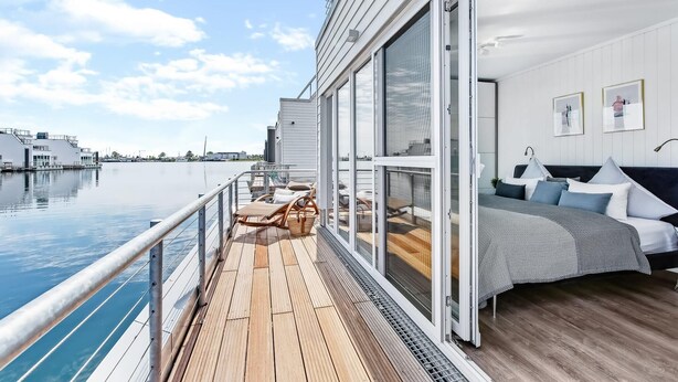 Una camera moderna con un'ampia porta a vetri scorrevoli che dà su un balcone con pavimento in legno e vista sull'acqua.