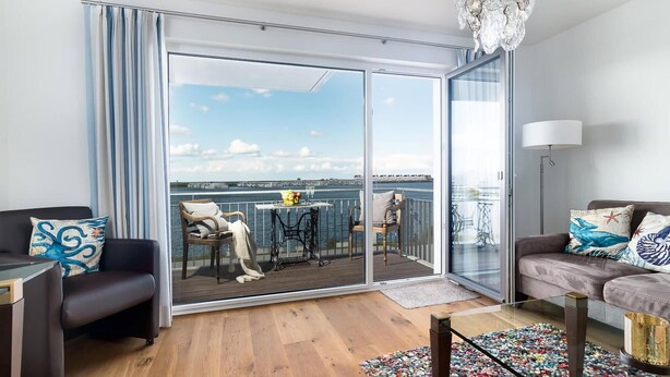 Una sala de estar moderna con un gran ventanal corredizo que da a un balcón con vista al agua y un paisaje urbano distante.