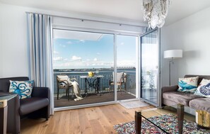 Un salon moderne doté d’une grande porte-fenêtre coulissante qui donne sur un balcon offrant une vue sur l’eau et sur le paysage urbain au loin.