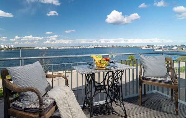 Un balcon avec deux chaises en bois, une table sur laquelle se trouve un panier de fruits et des verres, donnant sur un front de mer avec des bâtiments et des bateaux.