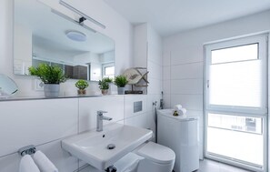 Une salle de bain moderne avec un lavabo blanc, des toilettes et un grand miroir. On trouve des plantes en pot sur le comptoir et une sculpture de poisson au-dessus du miroir.