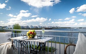 Une table avec un panier de fruits et un verre de vin blanc sur un balcon donnant sur une rivière, avec des bâtiments en arrière-plan.