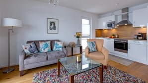 Un apartamento moderno con un sofá gris, una mesa de centro de vidrio, un sillón beige, una cocina con armarios blancos y una araña.