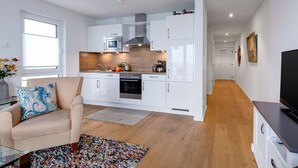 Una cocina moderna con armarios blancos, horno empotrado y microondas. Hay una zona de comedor con un sofá beige y una alfombra colorida.
