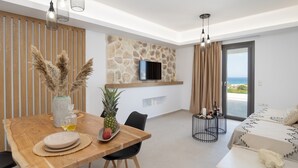 A modern living room with a stone wall, a flat-screen TV, a dining area with a wooden table, and a view of the sea through large windows. - BLUE SEA NAXOS  (Naxos)