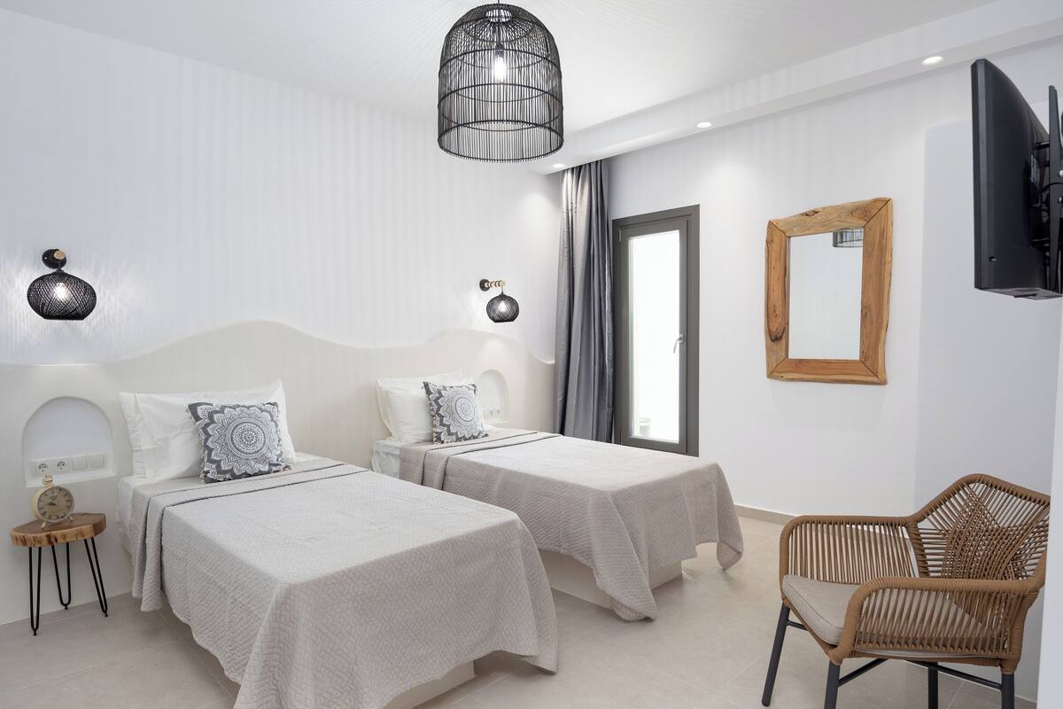 Quarto de hotel moderno com duas camas, uma cadeira de madeira, uma televisão e uma janela grande.
