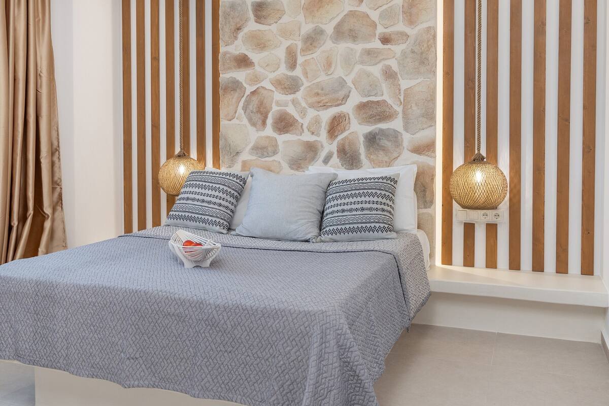 Quarto com parede de pedra, cama com almofadas estampadas e duas luminárias pendentes.
