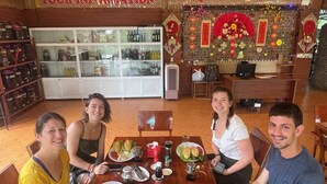 A group of people seated at a wooden table with plates of food, a glass cabinet with bottles, and a sign reading "TOUR INFORMATION". - HaGiang Eco Bungalow (Ha Giang 1)