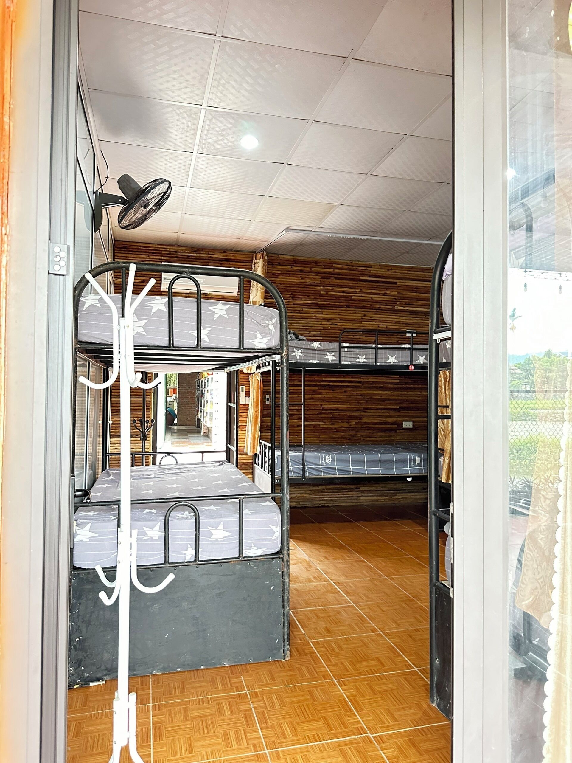 Una habitación con literas, ventilador de techo y ventana con vista a follaje.
