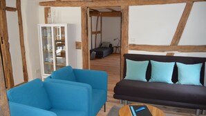 Una habitación con techo de madera, un sofá azul con cojines azul claro, un sofá negro, una mesa de centro de madera con un cuaderno y un aparador de vidrio con vajilla.