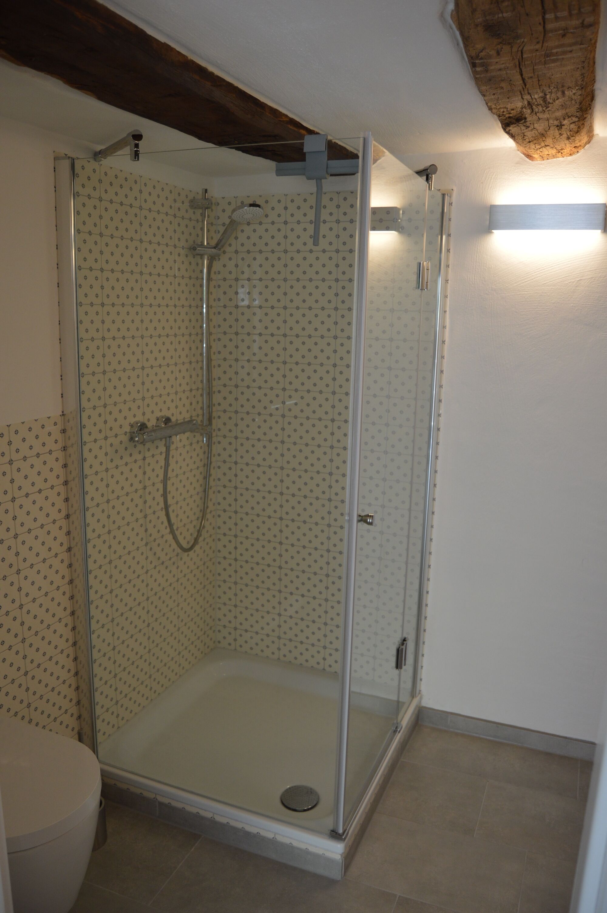 A glass-enclosed shower area with a tiled wall and a wooden ceiling beam.