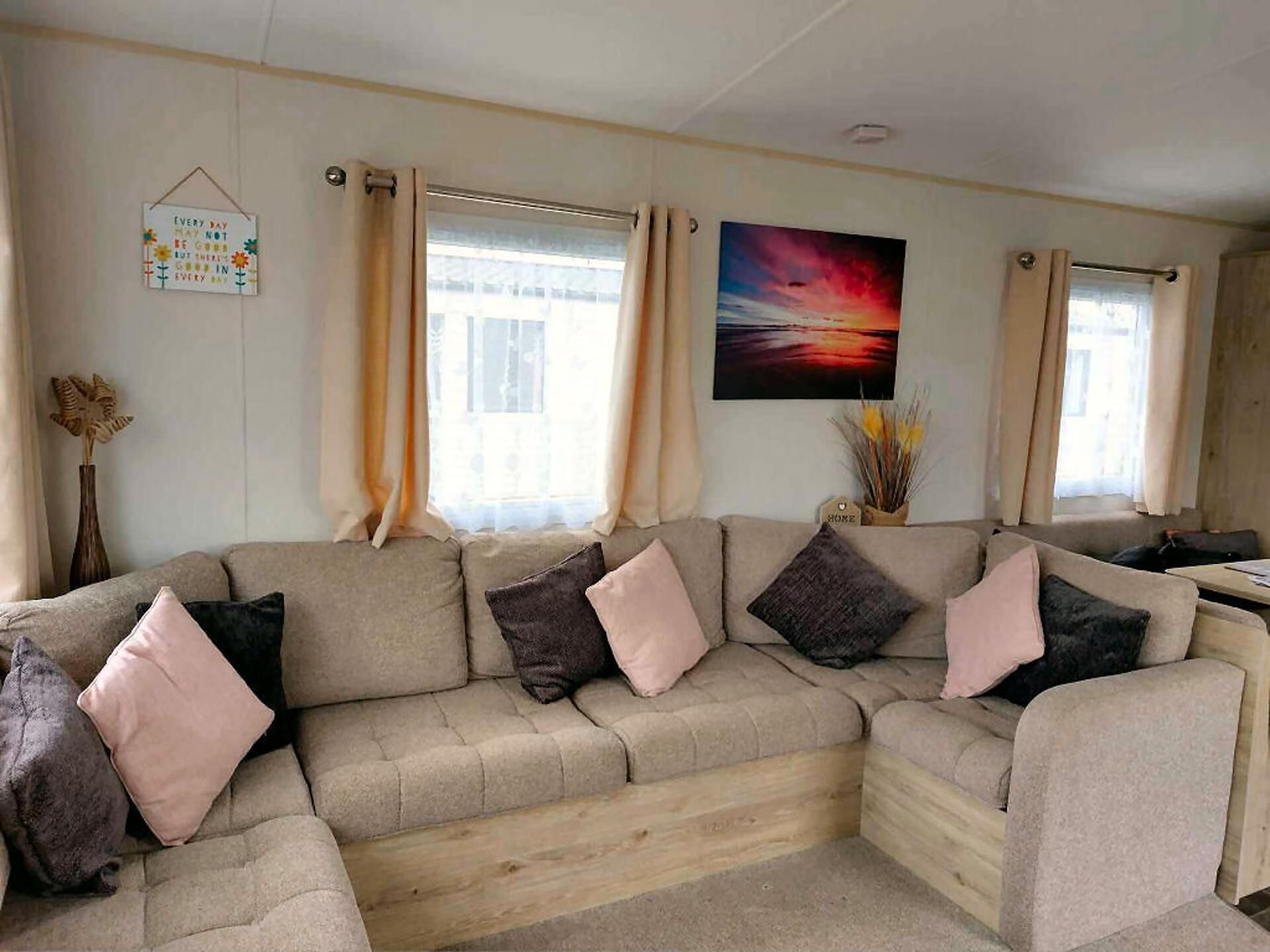 Un salon avec un canapé d’angle beige, un tableau représentant un coucher de soleil et un panneau accroché au mur.