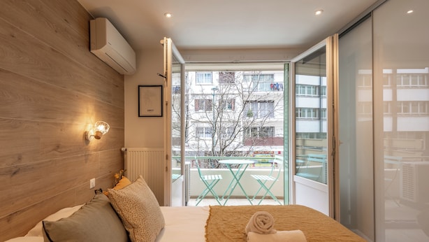 Un dormitorio moderno con cabecera de madera, una cama grande con una manta color beige, una mesita de noche con una silla y vistas al balcón exterior.