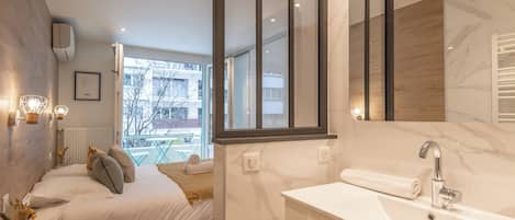 A modern bedroom with a large bed, bedside table with a vase, and a view of the outside.