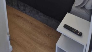 A small, simple room with a bed, a white nightstand, and a remote control on the nightstand.
