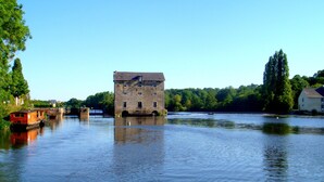 A historic stone building with a large window and a balcony, surrounded by calm water and greenery.
