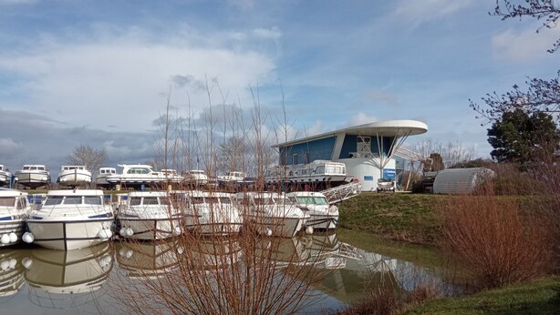 A marina with several boats docked, a large building with a unique curved roof, and a small body of water reflecting the boats. - Exclusive Stay on Triton 860 Fly (Pontailler-sur-Saône)