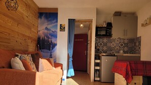 Un salon chaleureux avec un mur en bois, un canapé agrémenté de coussins à motifs, une cuisine équipée d’un four à micro-ondes et ornée de carreaux à motifs, et une porte donnant sur une autre pièce.
