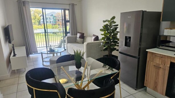 Een moderne keuken met een roestvrijstalen koelkast, een eethoek met zwarte stoelen en een glazen tafel, en een balkon met uitzicht op gebouwen en groen.