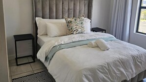 Een net opgemaakt bed met een grijs bekleed hoofdeinde, witte beddengoed en een kussen met een patroon.