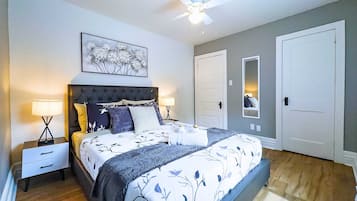 Une chambre à coucher comprenant un lit, des tables de chevet, un ventilateur de plafond, une œuvre d’art accrochée au mur et une porte.