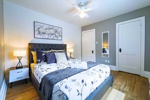 Une chambre à coucher comprenant un lit, des tables de chevet, un ventilateur de plafond, une œuvre d’art accrochée au mur et une porte.