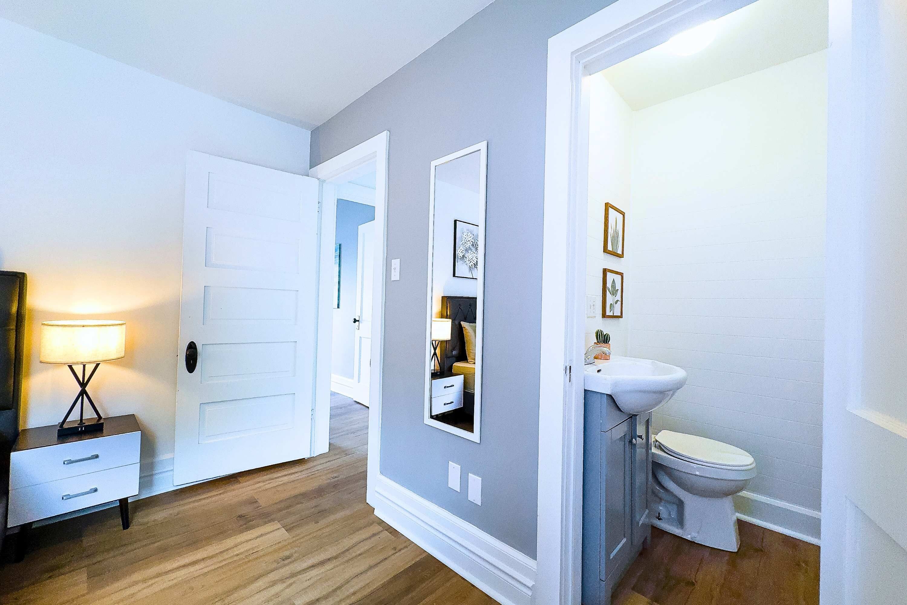 A hallway with a white door, a bedside table with a lamp, and a bathroom with a sink and toilet.