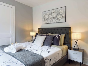 Un lit bien fait, recouvert d’une couvre-lit à motifs floraux, deux lampes de chevet, une table de chevet avec un tiroir et, au-dessus du lit, une œuvre d’art encadrée représentant des fleurs.