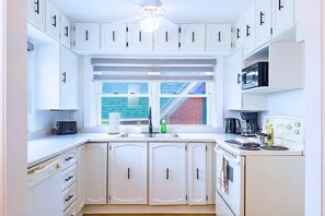 Une cuisine avec des meubles blancs, un four à micro-ondes encastré, une cuisinière et une fenêtre donnant sur un immeuble en briques.