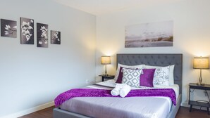 Une chambre moderne avec un lit recouvert d’un tissu gris, une literie aux motifs violets et blancs, et une tête de lit grise. On y trouve deux tables de chevet avec des lampes, une œuvre d’art accrochée au mur et un tableau floral encadré au-dessus du lit.