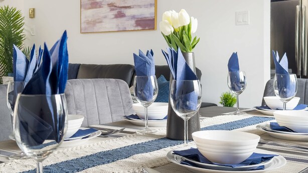 Un coin repas avec une table dressée pour quatre personnes, agrémenté de serviettes bleues, d’assiettes blanches et de verres à vin transparents.