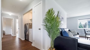 Un salon moderne avec un plancher en bois, un canapé noir orné de coussins à motifs, une plante en pot et une vue sur une buanderie.