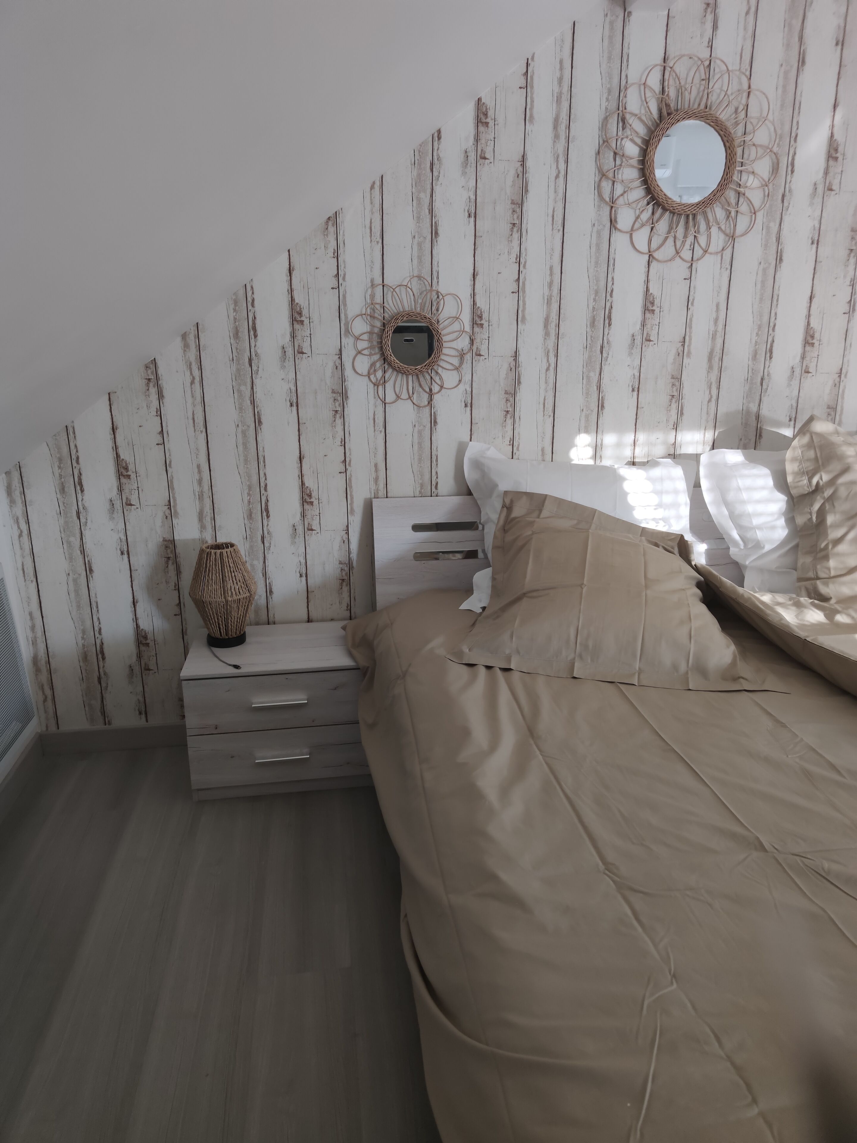 Ein Schlafzimmer mit schrägverdeckter Decke, einem Bett mit beiger Bettwäsche, einem Nachttisch mit Schubladen, einer Lampe und zwei an der Wand befestigten Spiegeln.