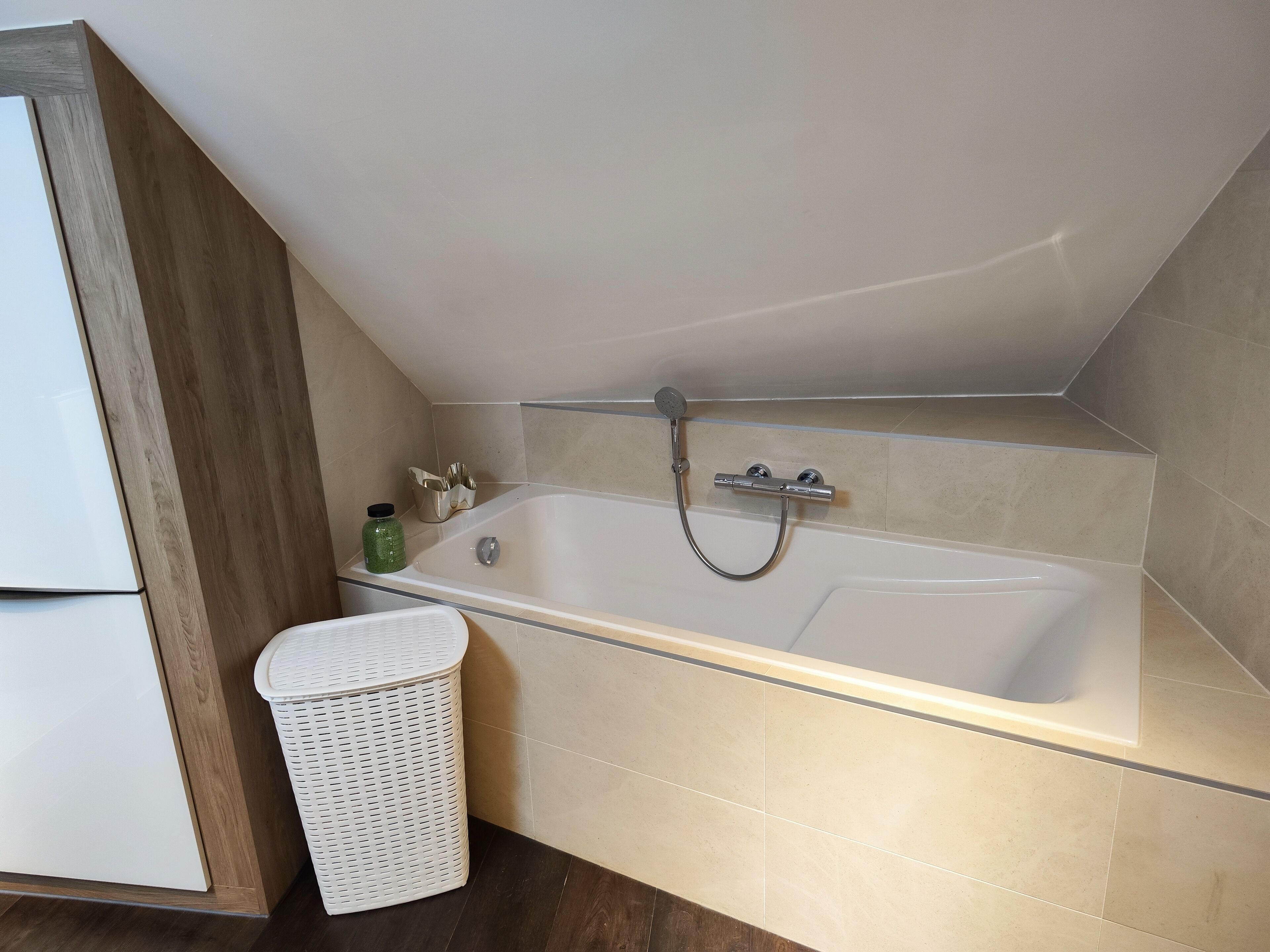 Une salle de bain avec une baignoire, une pomme de douche, un panier en osier blanc et une bouteille verte.