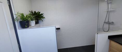 A modern bathroom with a shower, white tiled walls, a black tiled floor, and a white door. There are two potted plants on a shelf.