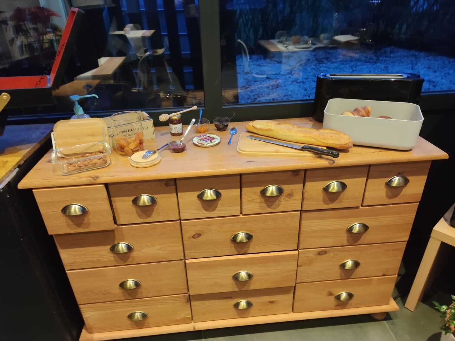 A wooden chest of drawers with multiple compartments, a wooden cutting board, a baguette, a honey dipper, jars of honey, and a jar of almonds.