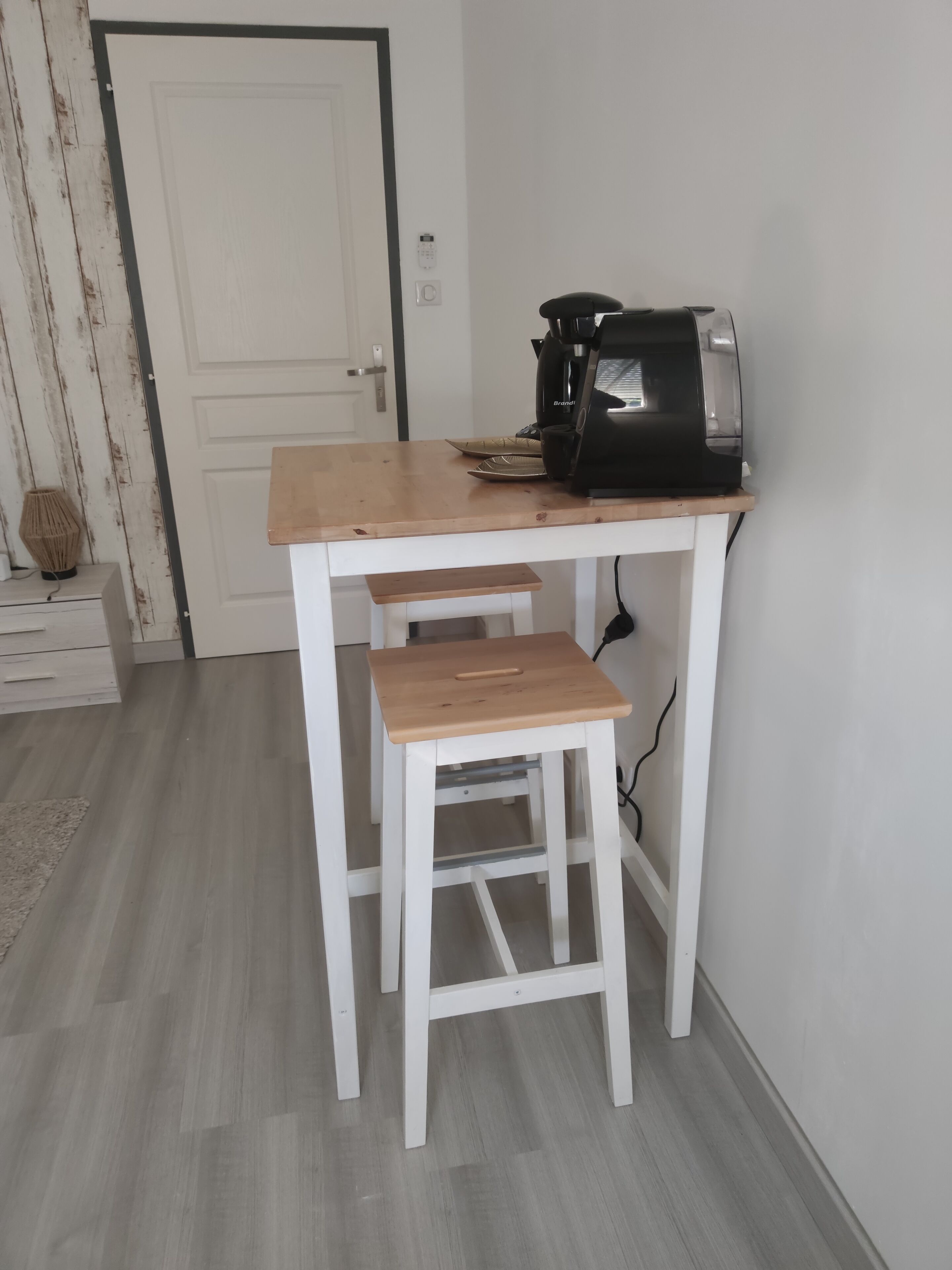 Una mesa pequeña de madera blanca con una cafetera encima, ubicada en una habitación con piso de madera clara y una puerta blanca al fondo.