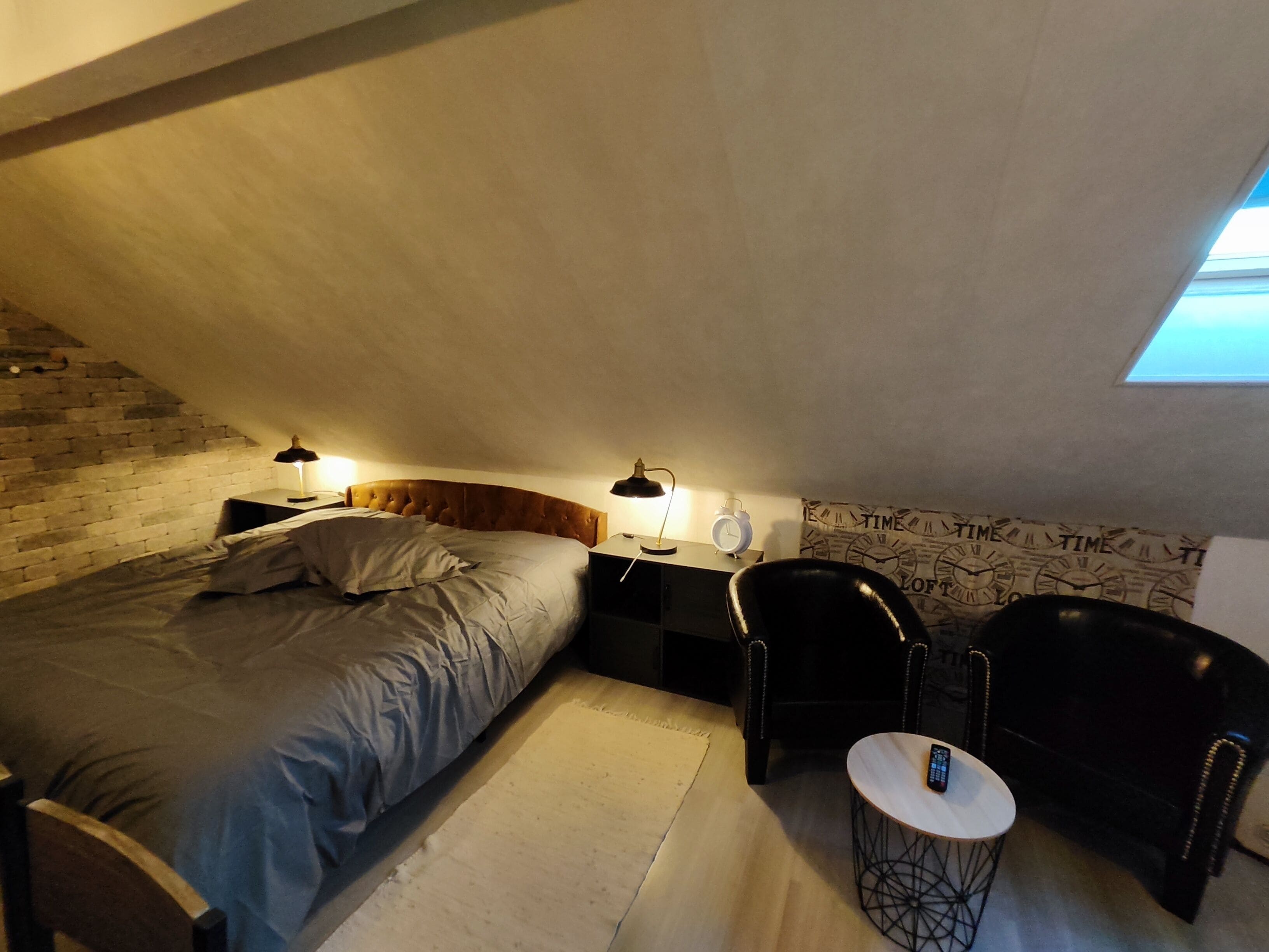 A cozy attic bedroom with a bed, two bedside tables with lamps, a chair, a small table, and a clock on the wall.