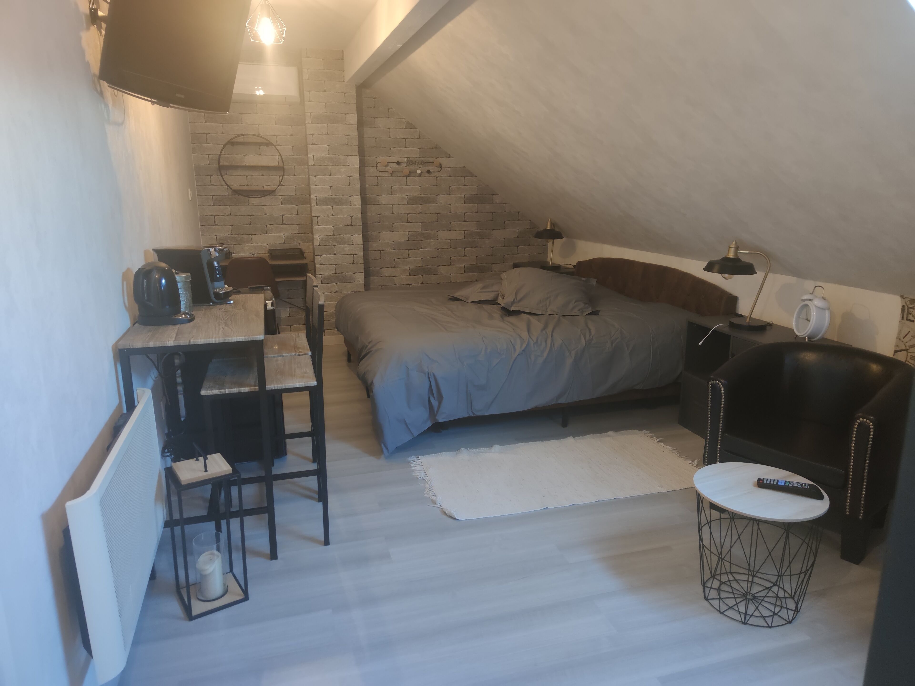 A compact, modern loft bedroom with a sloped ceiling, a bed with grey bedding, a desk with a chair, a small round table, and a black leather armchair.