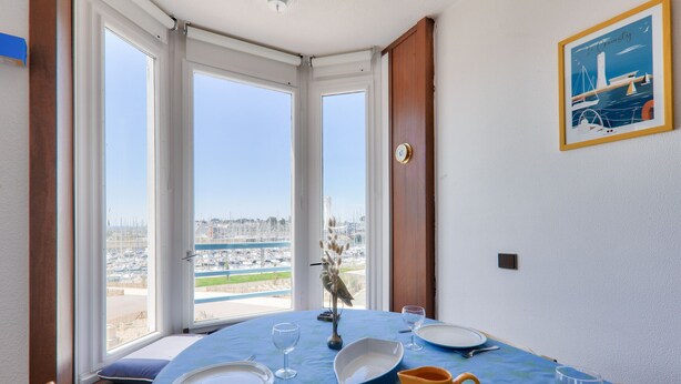 Un comedor con una mesa dispuesta para dos personas, ventanas amplias con vistas al puerto deportivo y un cuadro en la pared.