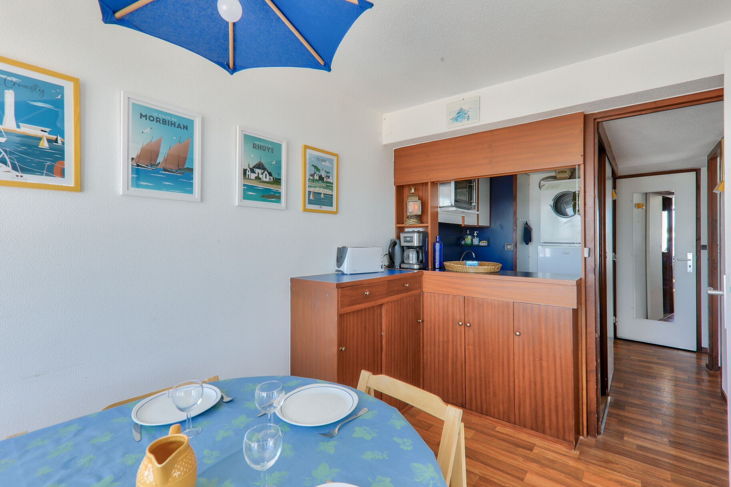 A dining area with a table set for two, a kitchen with wooden cabinets, and a blue umbrella overhead.