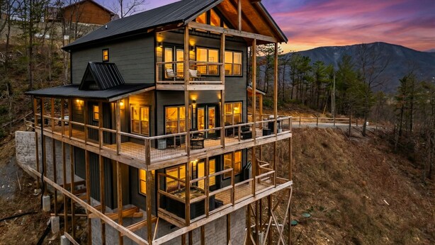 A two-story wooden cabin with a balcony, surrounded by trees and mountains.