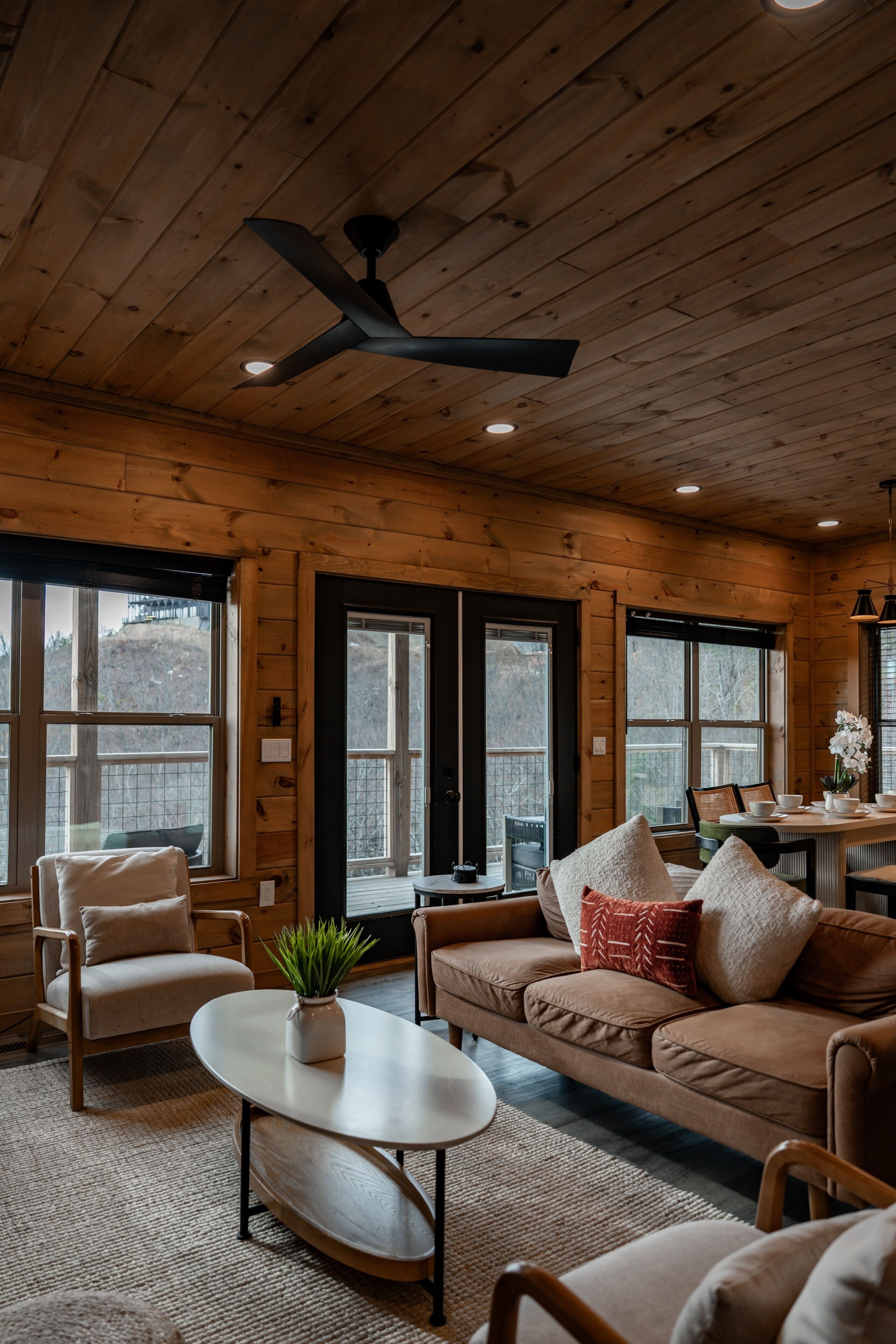 A cozy cabin interior with wooden walls, a ceiling fan, a dining table, and a sofa.