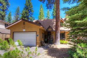 A two-story wooden cabin with a garage, surrounded by pine trees.
