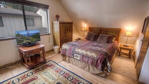 Un dormitorio con techo inclinado, una cama con ropa de cama estampada, dos lámparas de noche, un televisor sobre una mesita de madera y una alfombra con diseño tradicional.