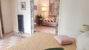 Quarto com cama, almofada verde, tapete estampado, radiador e vista para uma sala de estar com sofá e luminária.