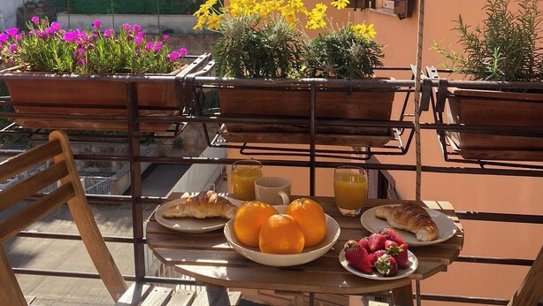 Uma área de refeições ao ar livre com uma mesa posta para o café da manhã, incluindo croissants, laranjas e morangos, com vista para um prédio e um jardim.