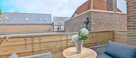 A rooftop terrace with a wooden fence, a round wooden table with a vase of flowers, and a wicker sofa.