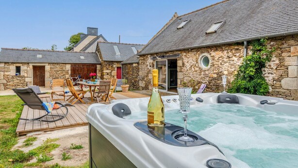 A hot tub with a bottle of champagne and a glass, surrounded by stone buildings and outdoor furniture. - Longère Charmante et Confortable, Proche de la Pointe Saint Mathieu (Plougonvelin)