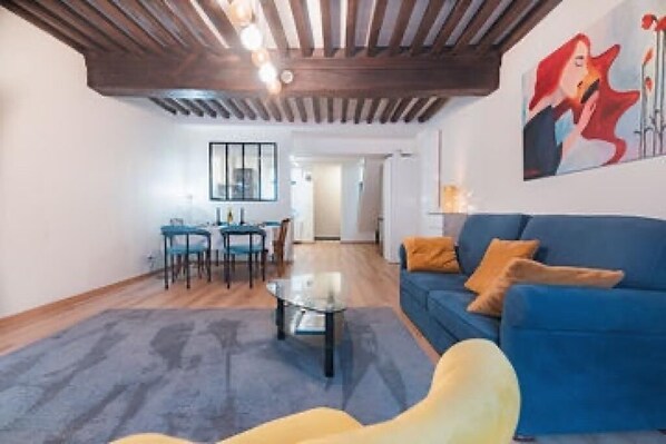 A living room with a blue sofa, a glass coffee table, and a dining area with chairs and a table. - Charming townhouse with a covered and furnished interior patio (Tournus)